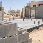 موقع بناء بيت في الكويت نهاراً تظهر فيه بلاطة خرسانية بمواسير بلاستيكية بارزة وجدران بلوك وعمال بخوذ أمان من بعيد