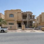 منزل سكني قديم في الكويت نهاراً بجانب منه جدران بلوك مكشوفة وسقالة منخفضة وعمال بخوذ أمان يظهرون من بعيد